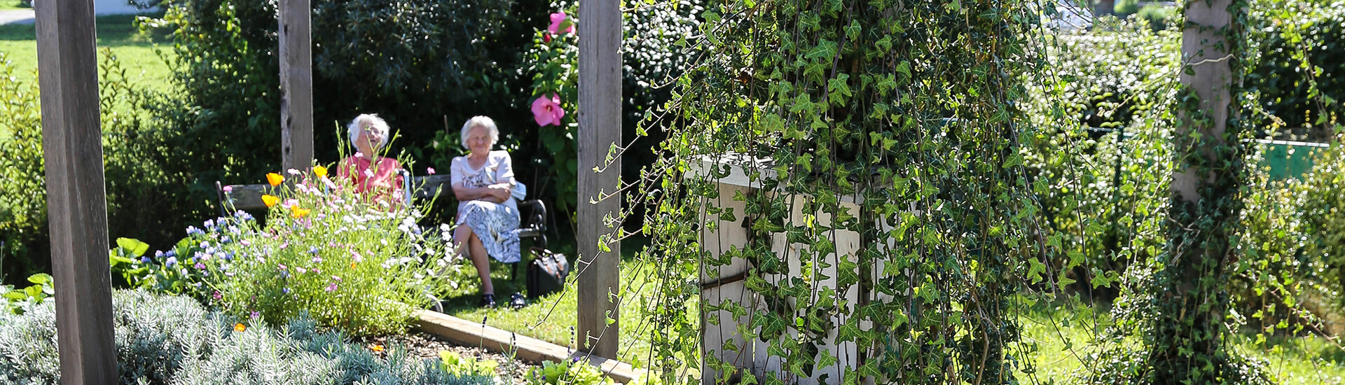 Zwei ältere Damen sitzen umgeben von Sträuchern und Blumen auf einer Holzbank im Garten.
